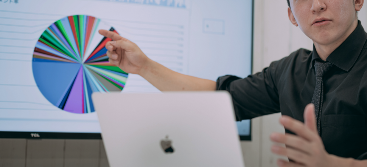 スーツ姿の男性がカラフルな円グラフを指しながらプレゼンをしている場面